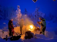 Vildmarksbyn Vildmarksliv i vinterlandskap under skymning. Tänd brasa med tre personer runtom.