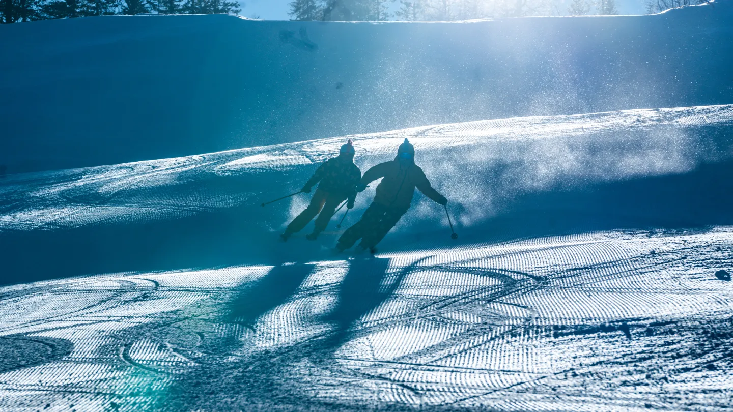 Färgbild - två skidåkare i skidbacke med solen i ryggen.