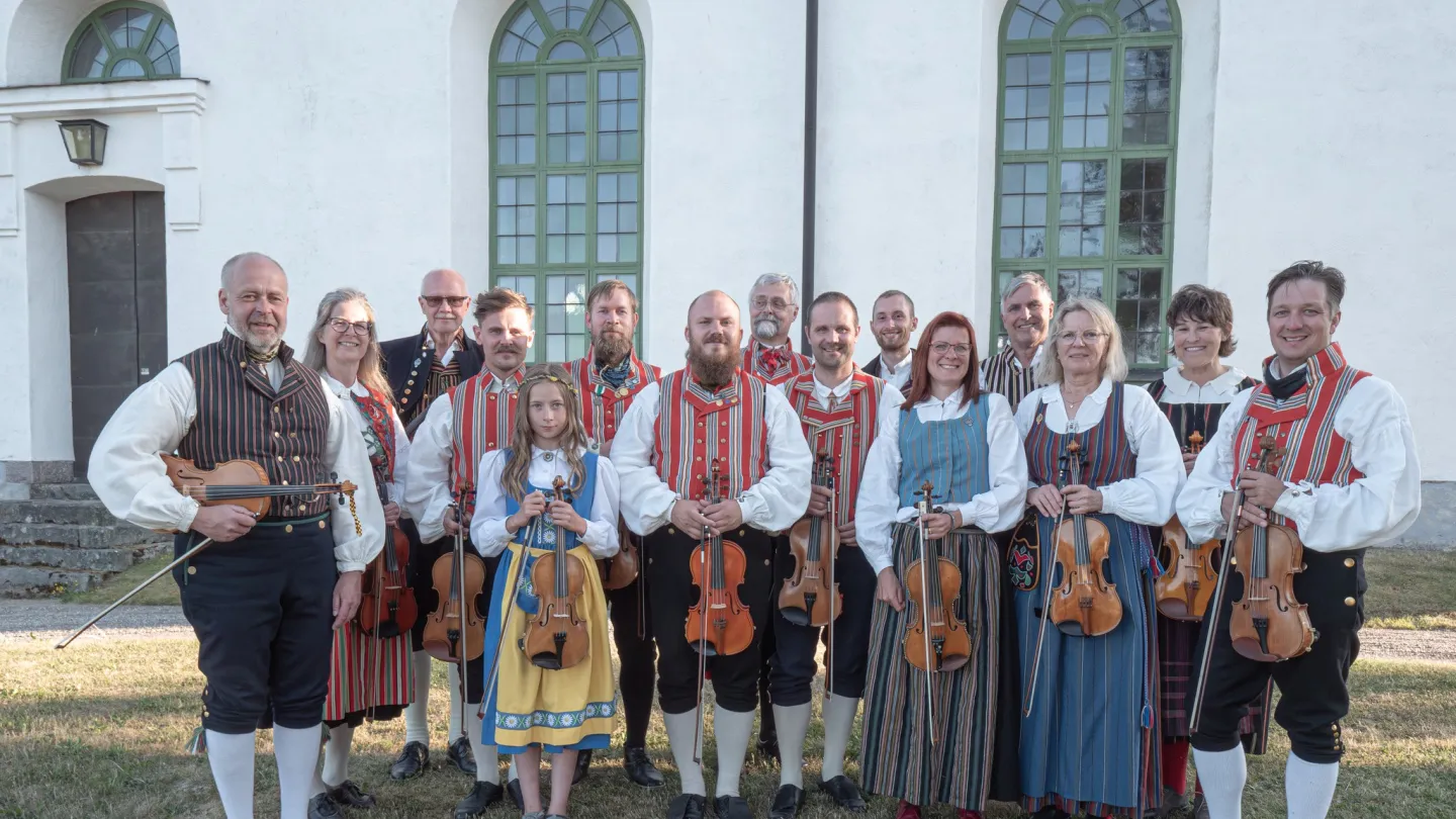 Färgbild utomhus - en grupp folkmusiker i folkdräkter framför kyrkfasad
