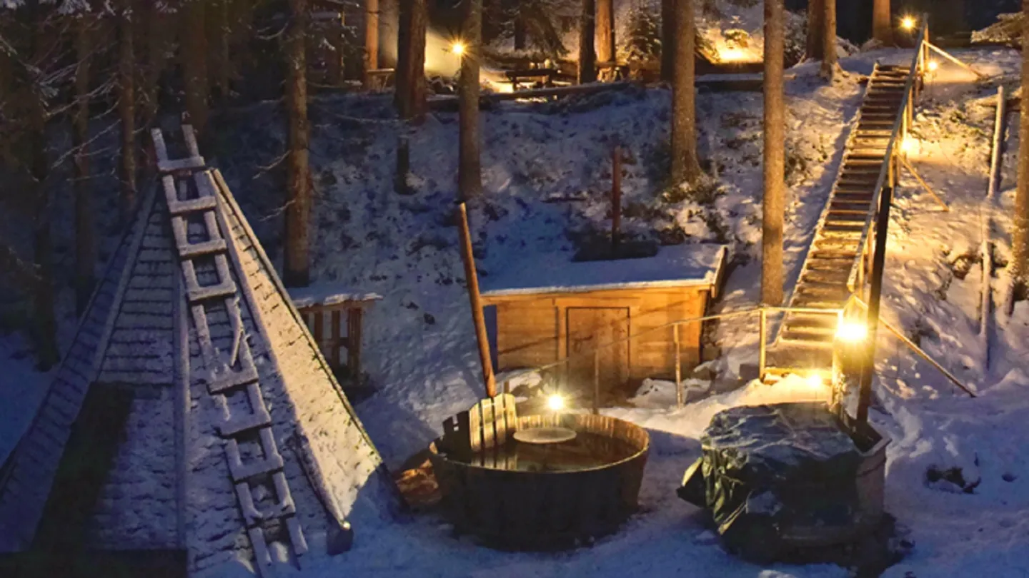Vildmarksby i vinterskrud i skymningen. Badtunna utomhus och träkåta.